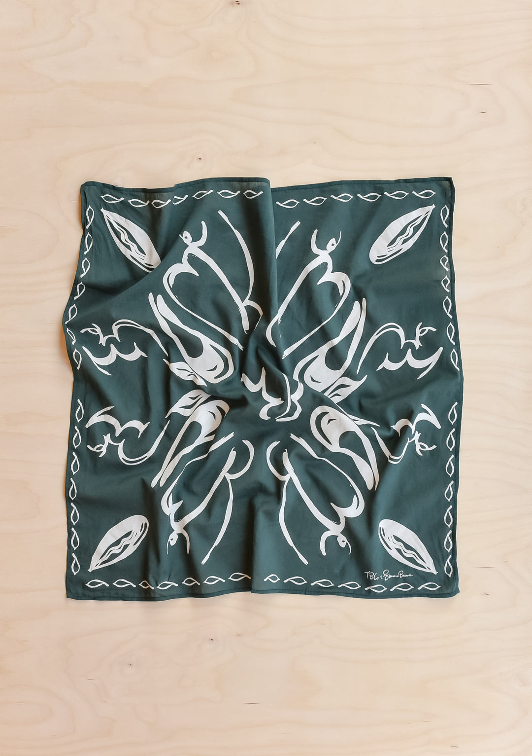The Artist Series TBCo x Simone Brewster Cotton Scarf, featuring a deep green with white leaf motifs, laid flat on a wood surface.