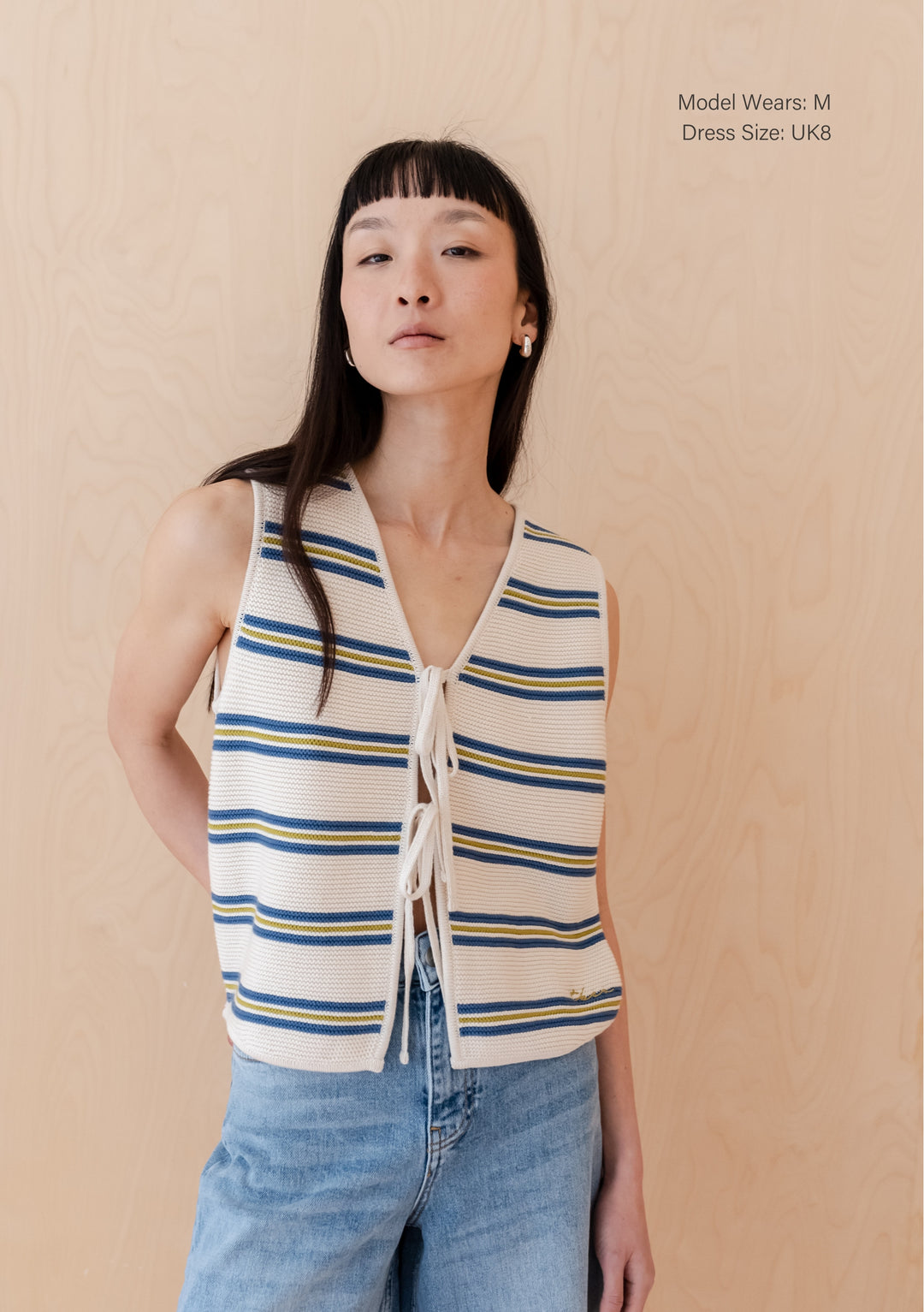 A person wearing TBCo Cotton Knitted Vest in Blue Stripe with adjustable ties, paired with light-wash denim against beige background
