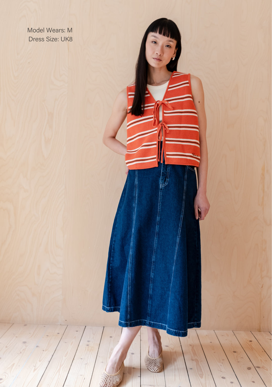 A person wearing TBCo Cotton Knitted Vest in Tangerine Stripe with front ties, paired with denim midi skirt and woven shoes