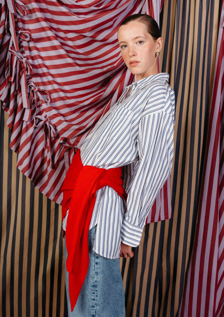 Model wearing a large merino triangle scarf in red by TBCo tied around waist over striped shirt and denim skirt