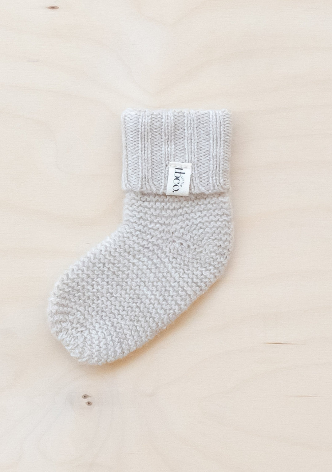 TBCo Merino Wool Baby Socks in Oatmeal Melange shown flat on wooden surface, displaying ribbed cuff and knitted texture