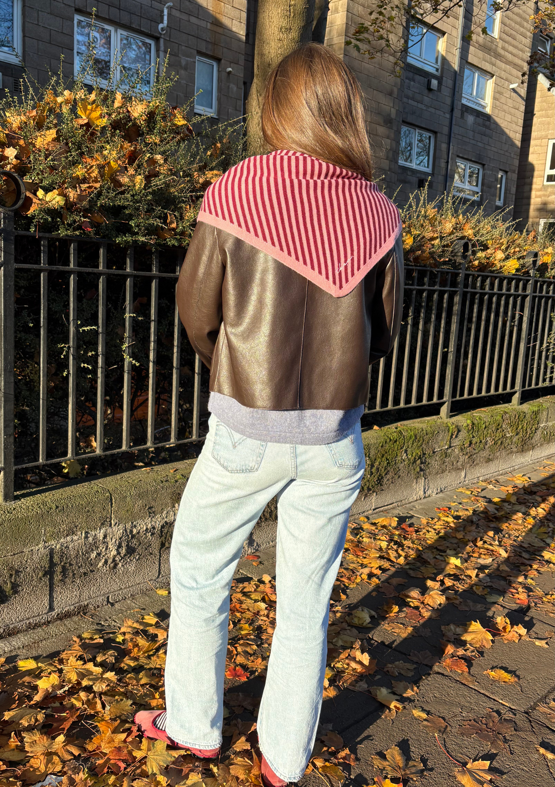 Woman outdoors wearing a Large Merino Triangle Scarf in Pink Stripe by TBCo with autumn leaves on the ground