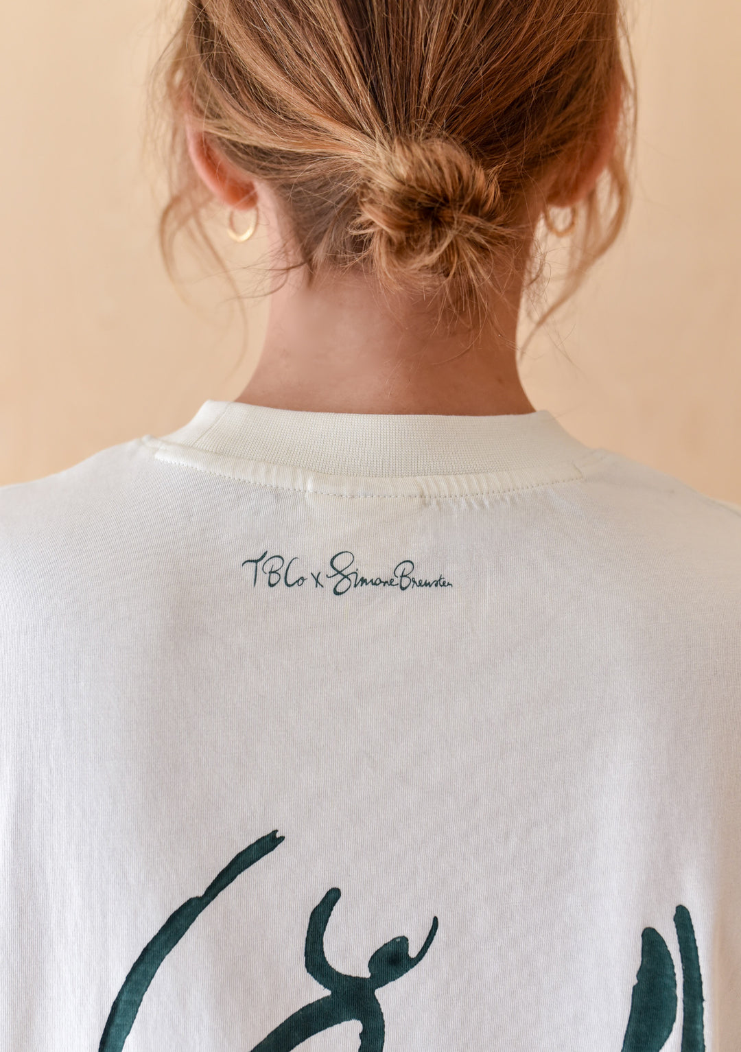 Back view of The Artist Series TBCo x Simone Brewster Cotton T-Shirt, displaying a green brushstroke graphic and script logo, worn with gold hoop earrings.