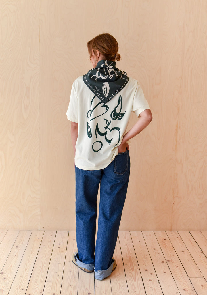 Person wearing The Artist Series TBCo x Simone Brewster Cotton T-Shirt with abstract brushstroke design, relaxed denim jeans, bandana, and sandals, against a wood-paneled backdrop.