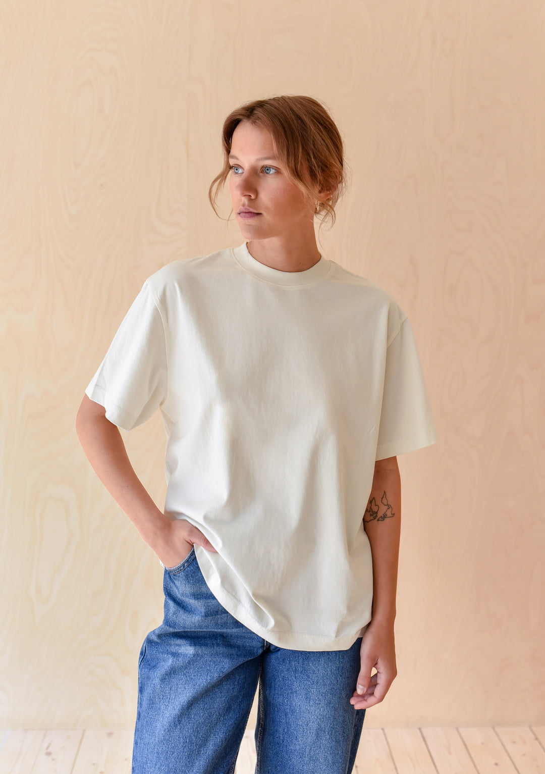 Young woman wearing The Artist Series TBCo x Simone Brewster Cotton T-Shirt, off-white, short-sleeve, paired with blue jeans, showcasing a minimalist, artistic design.