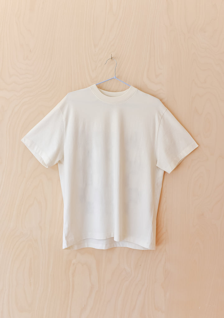 The Artist Series TBCo x Simone Brewster Cotton T-Shirt, plain white short-sleeve, on metal hanger against wood-grain wall, showcasing sustainable artistry in fashion.