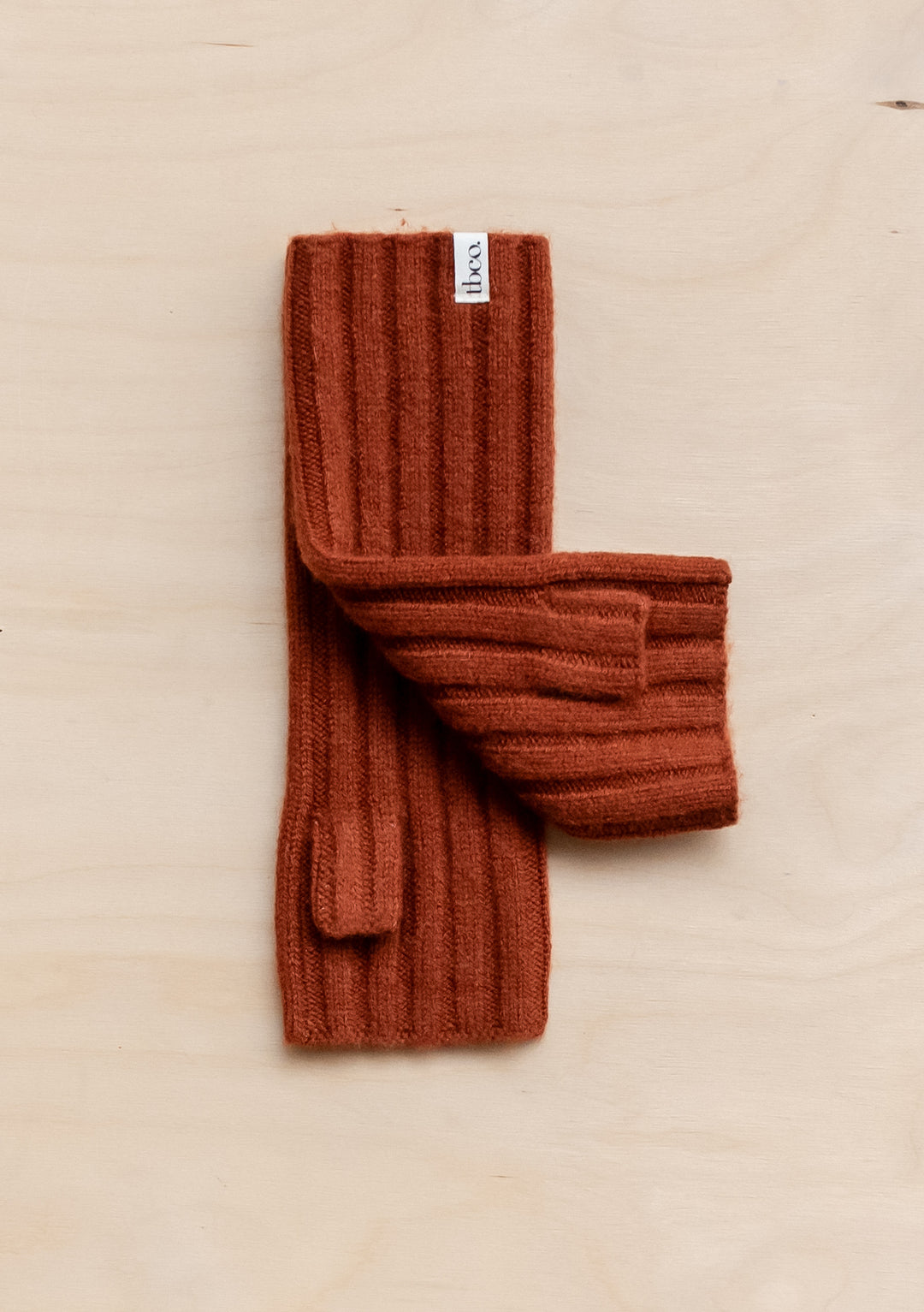 TBCo Cashmere & Merino Wrist Warmers in rust colour, ribbed knit pattern, displayed flat on beige surface, showing cosy winter accessories