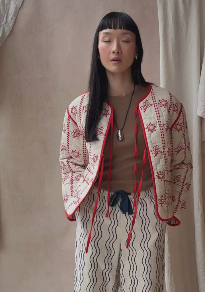 A model poses against a neutral ply backdrop, wearing a TBCo quilted jacket in cream and red, with a brown t-shirt underneath and TBCo cream and blue wave pattern trousers.