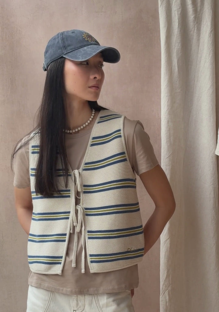 A model poses against a neutral ply backdrop, wearing a TBCo cream and blue striped vest waistcoat with a grey t-shirt underneath and blue baseball cap.