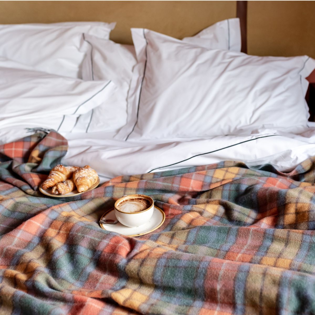 TBCo Recycled Wool Buchanan Antique Tartan Blanket in a rich check pattern draped over a cosy bed with crisp white linens. A warm and inviting bedroom setting featuring a cup of coffee and croissants, perfect for a relaxed morning