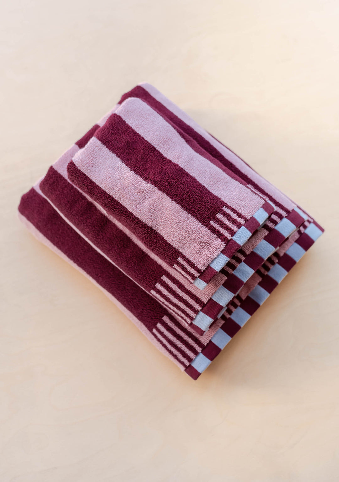 Stack of TBCo Terry Cotton Towels in Burgundy Stripe displaying plush texture and blue checkerboard edge detail on wood surface
