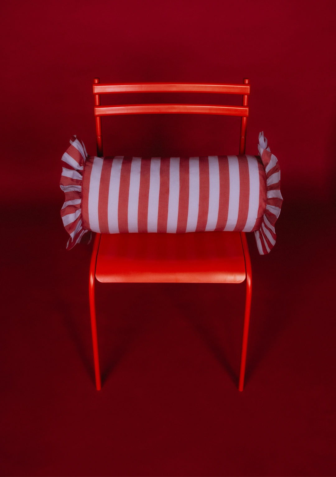 Bright-red chair with a TBCo Cotton Cushion Cover in Lilac Stripe, bolster-style, featuring red-and-white vertical stripes and ruffled ends, designed in Edinburgh.