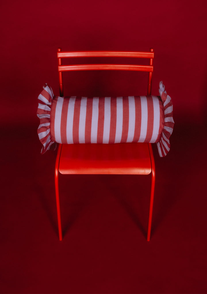 Bright-red chair with a TBCo Cotton Cushion Cover in Lilac Stripe, bolster-style, featuring red-and-white vertical stripes and ruffled ends, designed in Edinburgh.