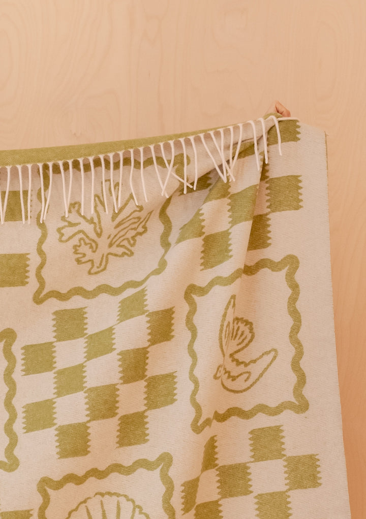TBCo Lambswool Blanket in Green Motif with decorative leaf and shell patterns and delicate fringed edges against pale wood background