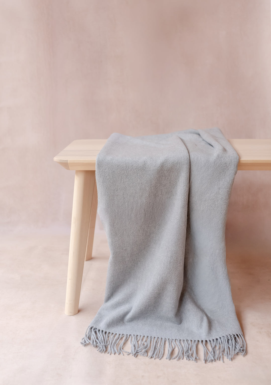 Cashmere Small Blanket in Light Grey Melange with fringe draped over bench, soft woven fabric by TBCo luxury collection