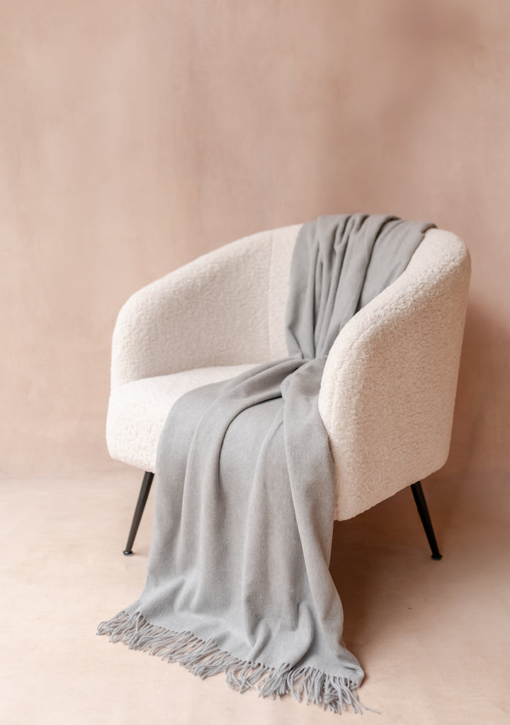 TBCo Cashmere Blanket in Light Grey Melange with fringe draped over a modern cream armchair in soft natural light