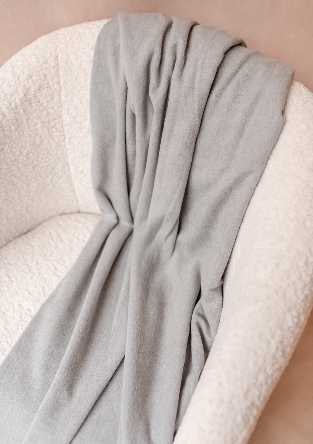 TBCo Cashmere Blanket in Light Grey Melange draped over a cream textured chair showcasing its soft luxurious fabric.