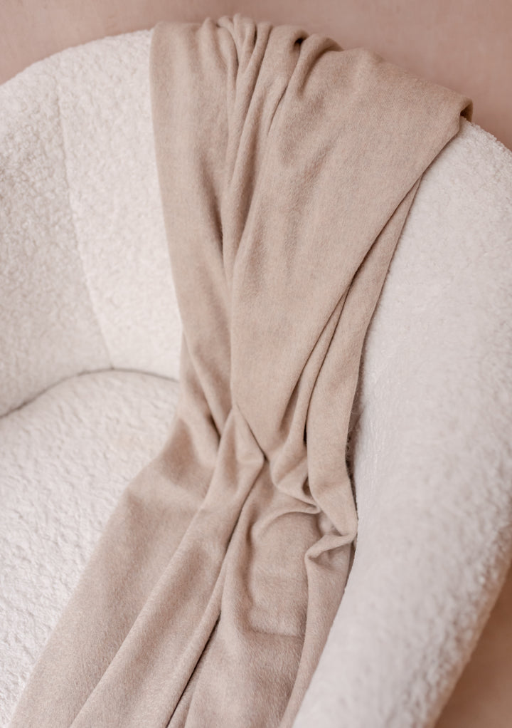 Cashmere Blanket in Oatmeal Melange by TBCo draped over a white boucle armchair showcasing its soft texture and warmth