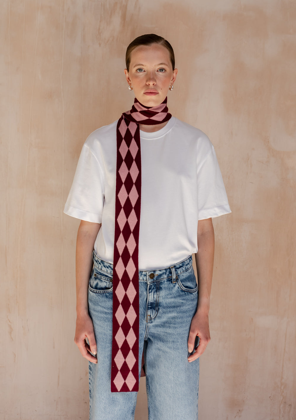 Model wearing a TBCo Merino Wool Skinny Scarf in Burgundy Harlequin elegantly draped around her neck. with a white t-shirt and white jeans.