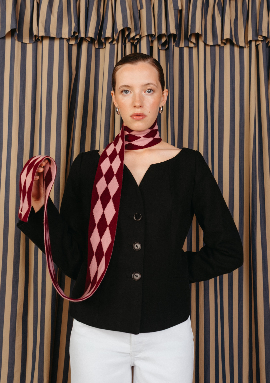 Model wearing a fitted black jacket and white pants, showcasing a TBCo Merino Wool Skinny Scarf in Burgundy Harlequin, elegantly draped around her neck.