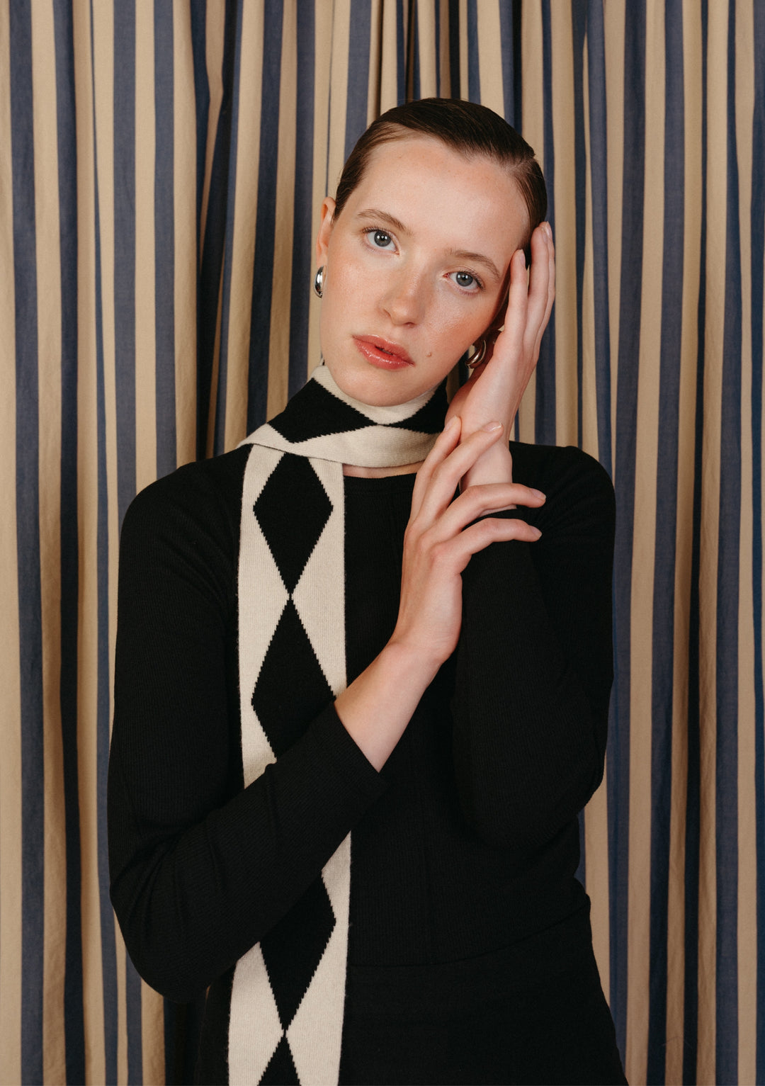 Young woman wearing Merino Wool Skinny Scarf in Black Harlequin pattern, styled with a black top, silver hoop earrings, against a muted striped backdrop.