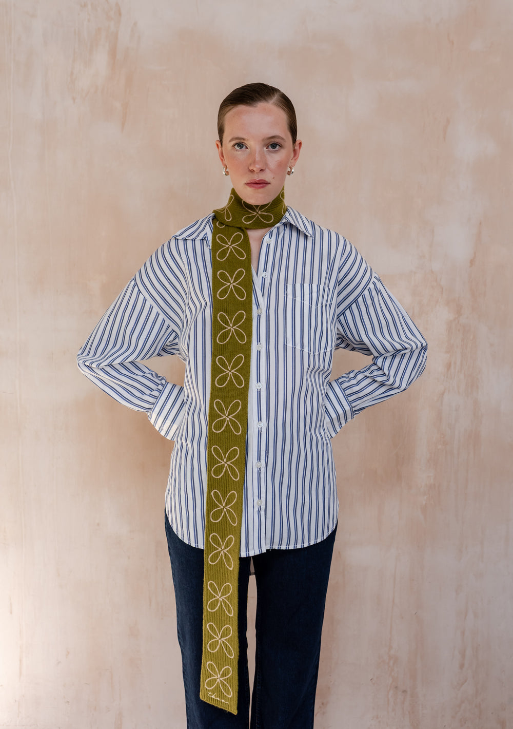 Model wearing a TBCo Merino Wool Skinny Scarf in Green Flora, adorned with white floral embroidery, layered over a blue stripe shirt, and dark jeans.