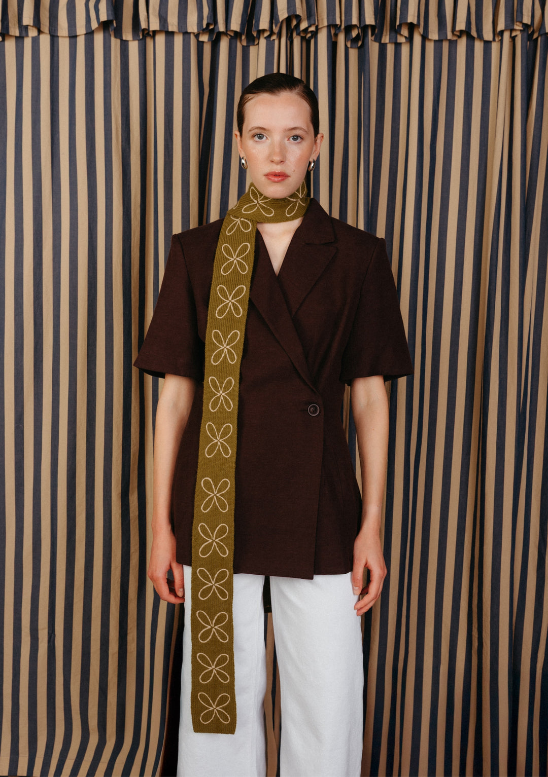 Model wearing a TBCo Merino Wool Skinny Scarf in Green Flora, adorned with white floral embroidery, layered over black shirt and white jeans in front of stripe curtains