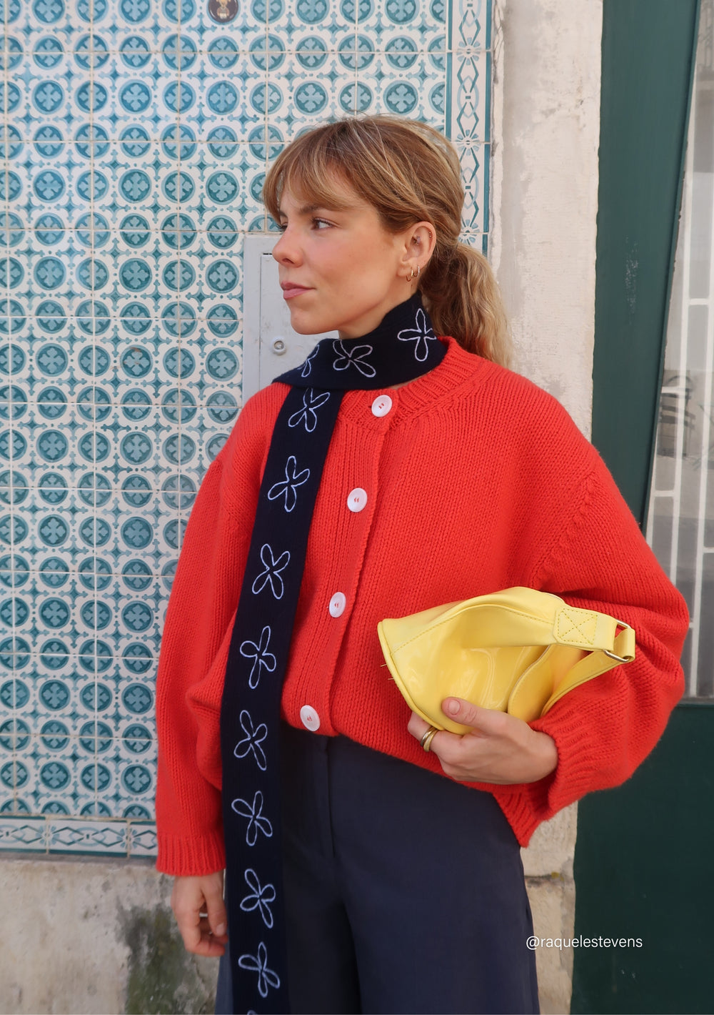 Model wearing a TBCo Merino Wool Skinny Scarf in Navy Flora, adorned with white floral embroidery, layered over a red cardigan, holding a yellow pouch.