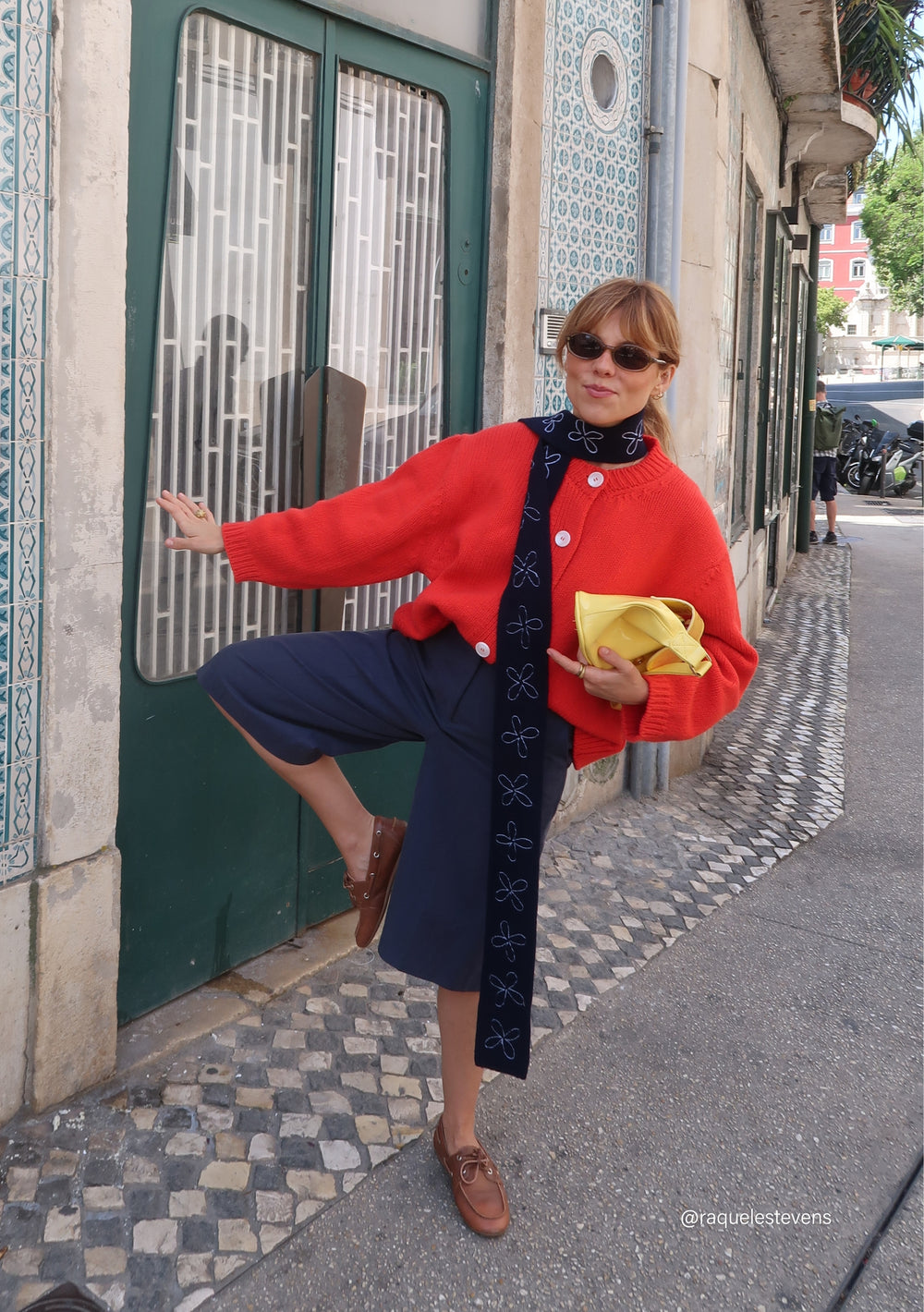 Model wearing a TBCo Merino Wool Skinny Scarf in Navy Flora, adorned with white floral embroidery, layered over a red cardigan, holding a yellow pouch.