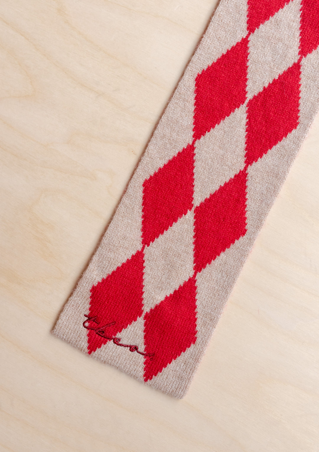 Close-up of TBCo Merino Wool Skinny Scarf in Red Harlequin showing red and oatmeal diamond pattern on wooden surface