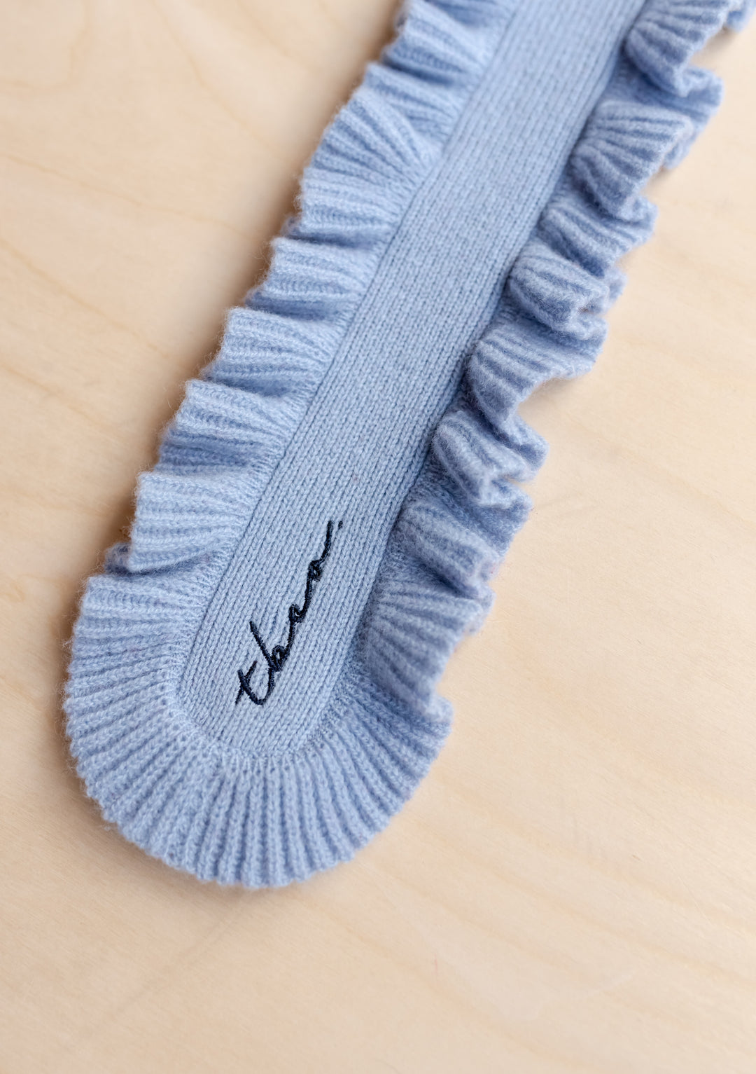 Close-up of TBCo Merino Wool Skinny Scarf in Blue Ruffle showing soft texture and delicate ruffled trim on light wood surface