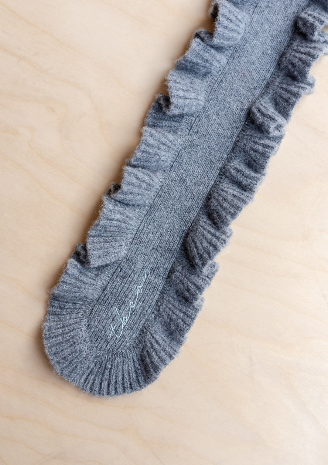Close-up of TBCo Merino Wool Skinny Scarf in Grey Ruffle showing soft texture and delicate ruffled trim on light wood background