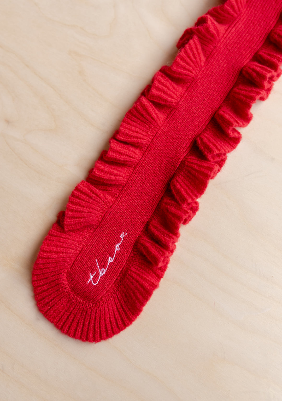 Close-up of TBCo Merino Wool Skinny Scarf in Red Ruffle showing ribbed texture and embroidered detail on light wood background