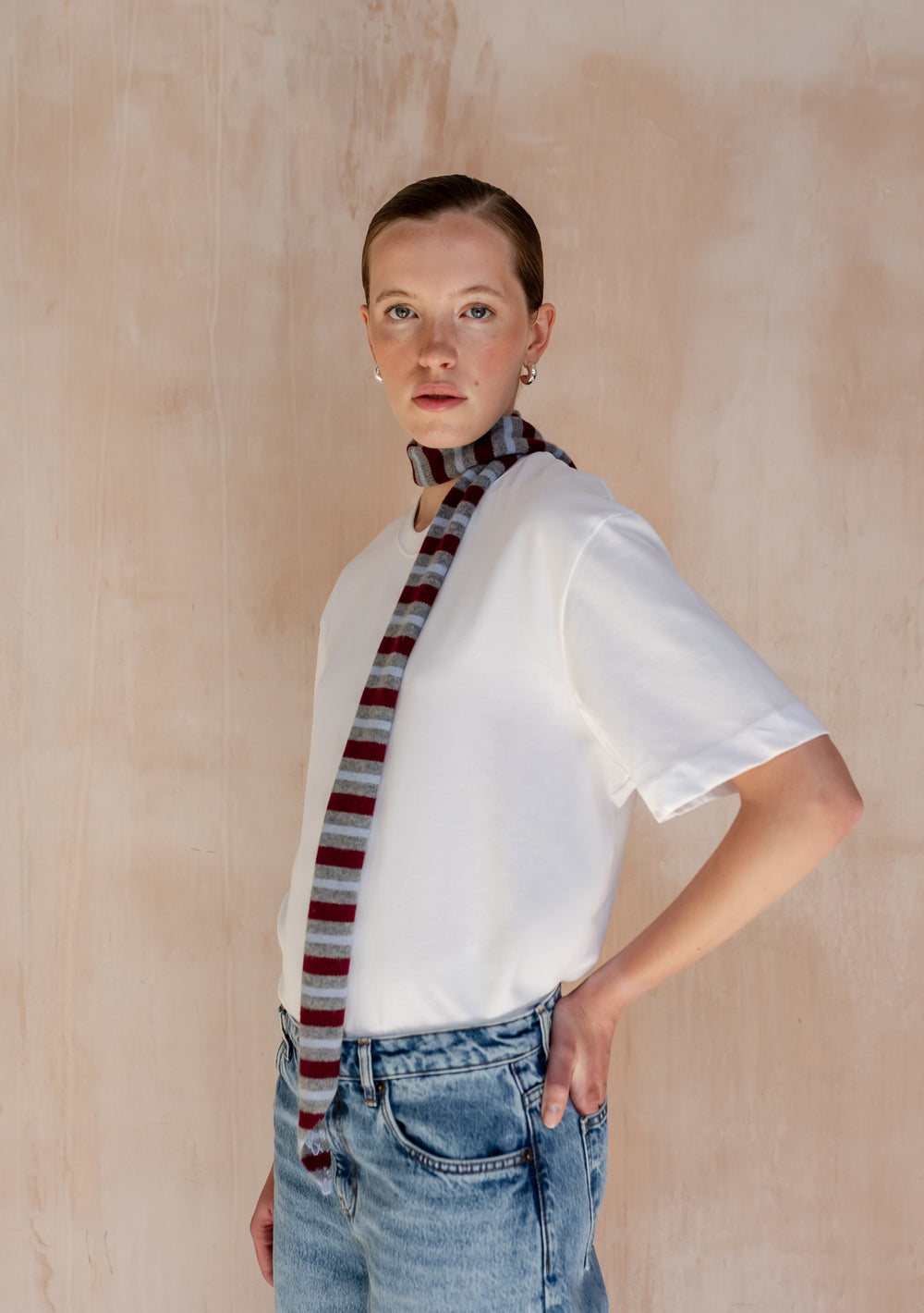 Model wearing a TBCo Merino Wool Neck Tie in Burgundy Stripe, styled over a white T-shirt and light-wash jeans, showcasing its subtle elegance and transitional-inspired design.