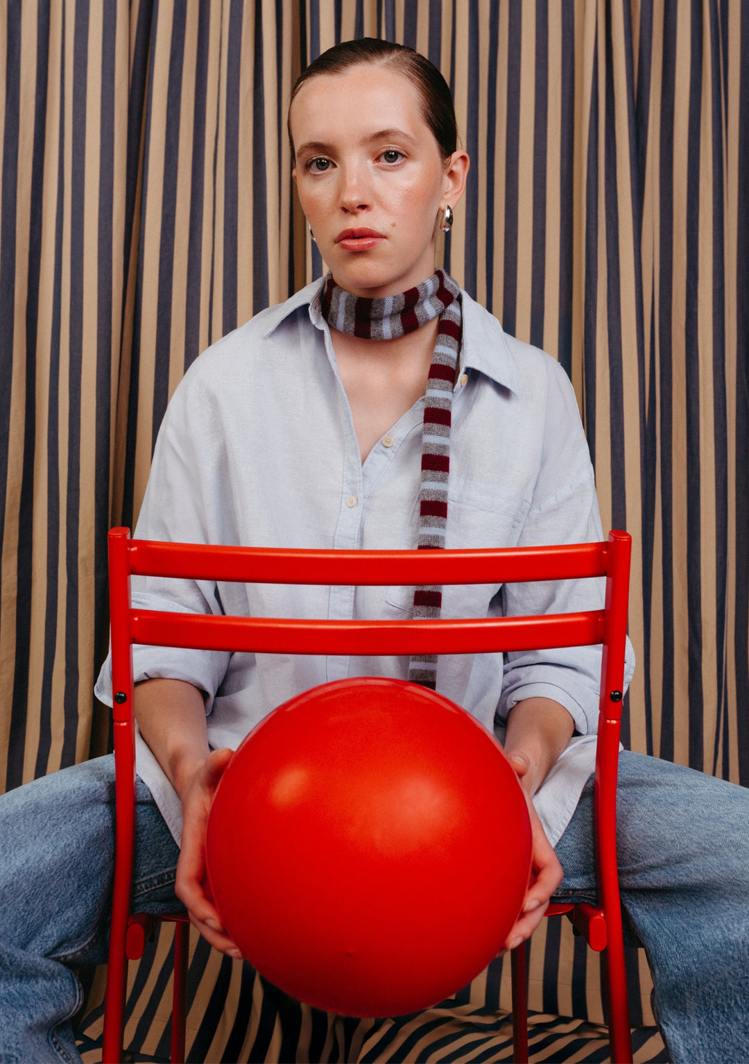 Model wearing a TBCo Merino Wool Neck Tie in Burgundy Stripe, styled with a blue shirt and blue jeans sitting on a red chair holding a red ball