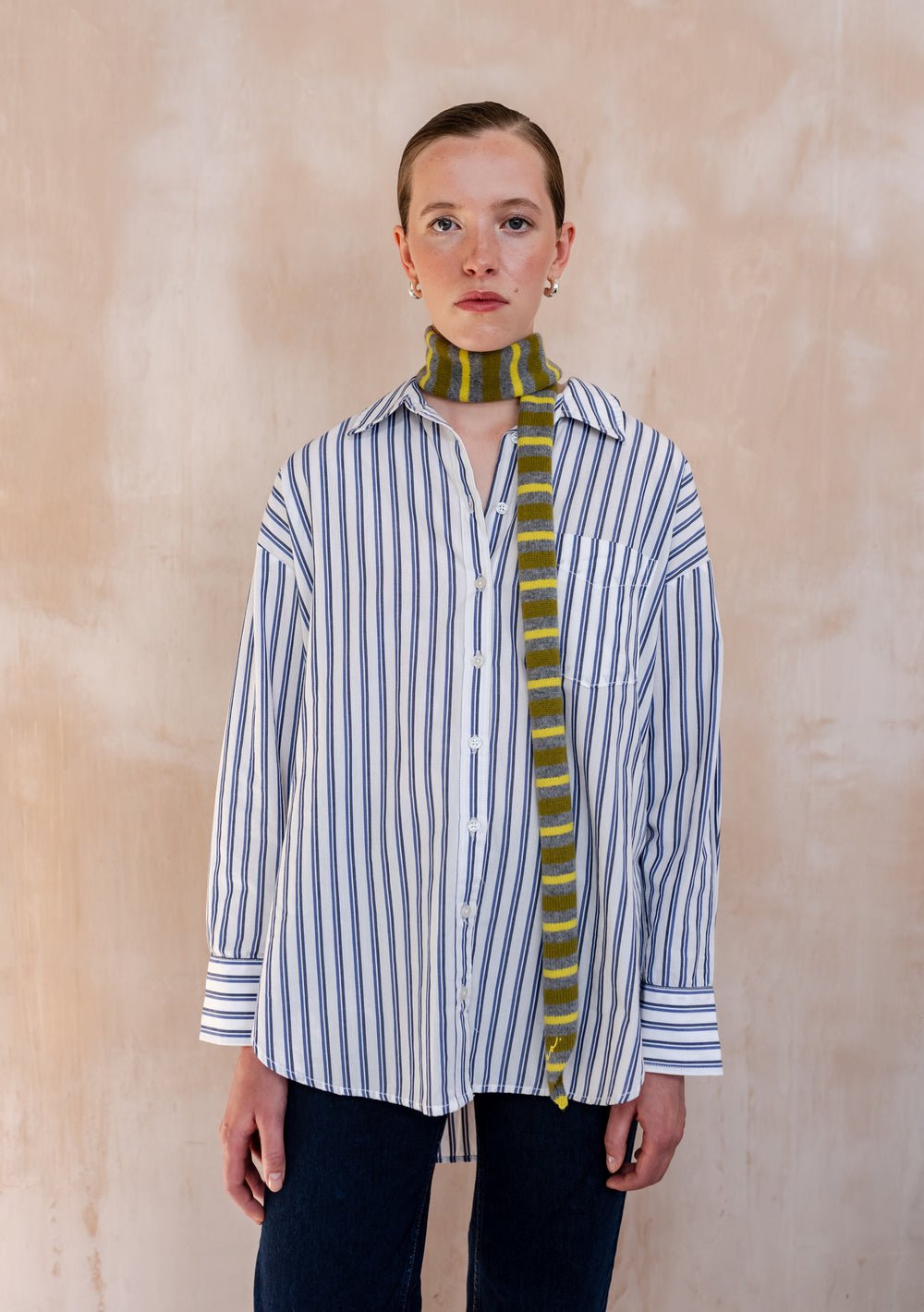 Model wearing a TBCo Merino Wool Neck Tie in Green Stripe, styled over a blue stripe shirt and dark jeans, showcasing its subtle elegance and transitional-inspired design.