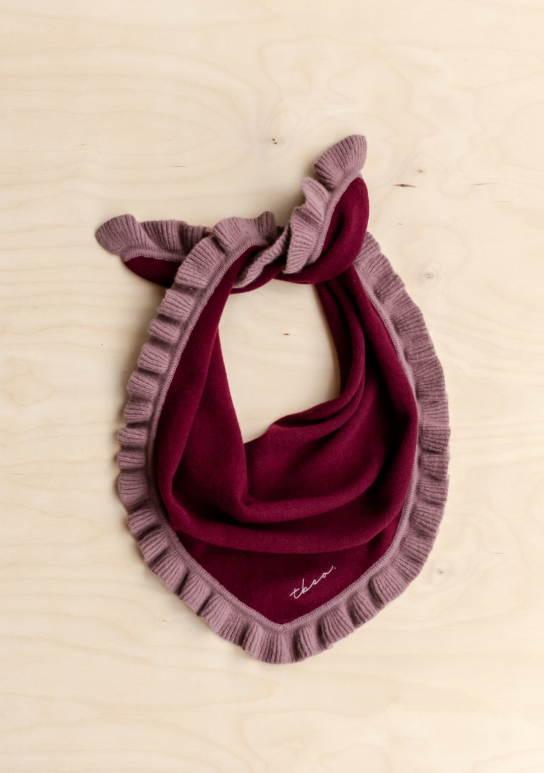 TBCo Merino Wool Triangle Scarf in Burgundy Ruffle, featuring a burgundy knit with pink ruffled edges, displayed on a wooden surface, showcasing its soft texture and elegant design.