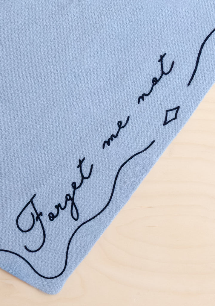 Close-up of TBCo Merino Wool Triangle Scarf in Forget Me Not showing pale blue fabric with embroidered text detail