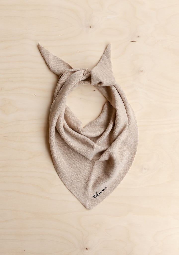 TBCo Merino Wool Triangle Scarf in Oatmeal, featuring taupe knit, black embroidery, and knotted ends, laid flat on wooden surface.