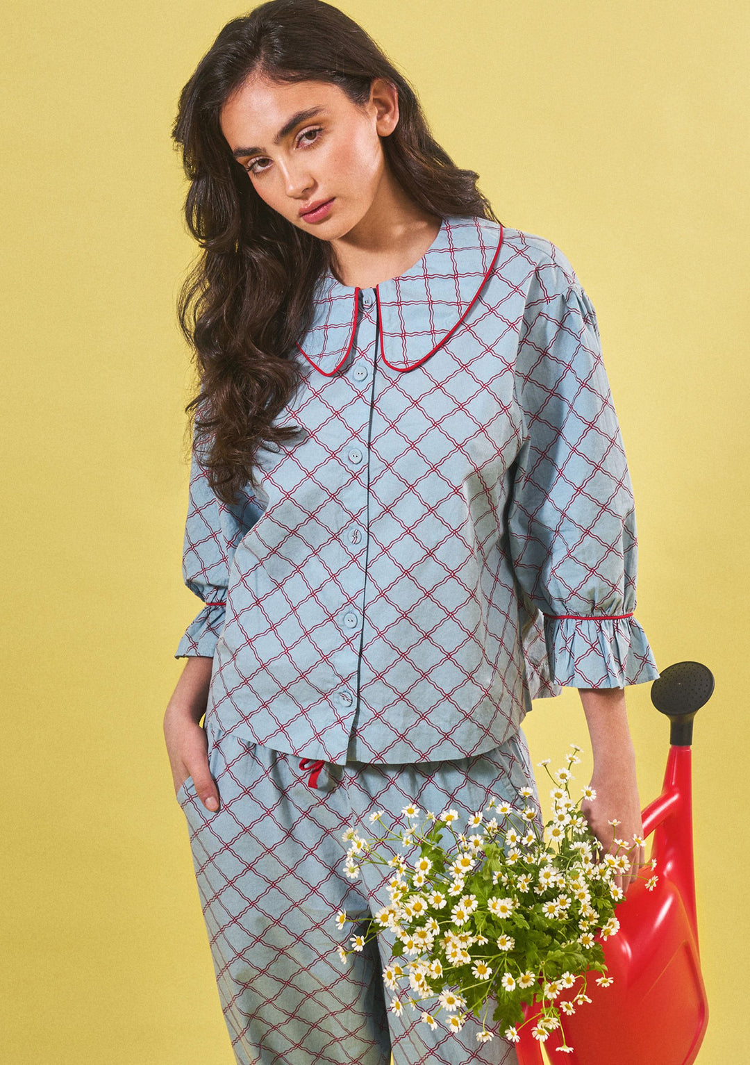 Model wearing TBCo Cropped Cotton Pyjamas in Blue Lattice, second gallery image, holding red watering can, portrait view.