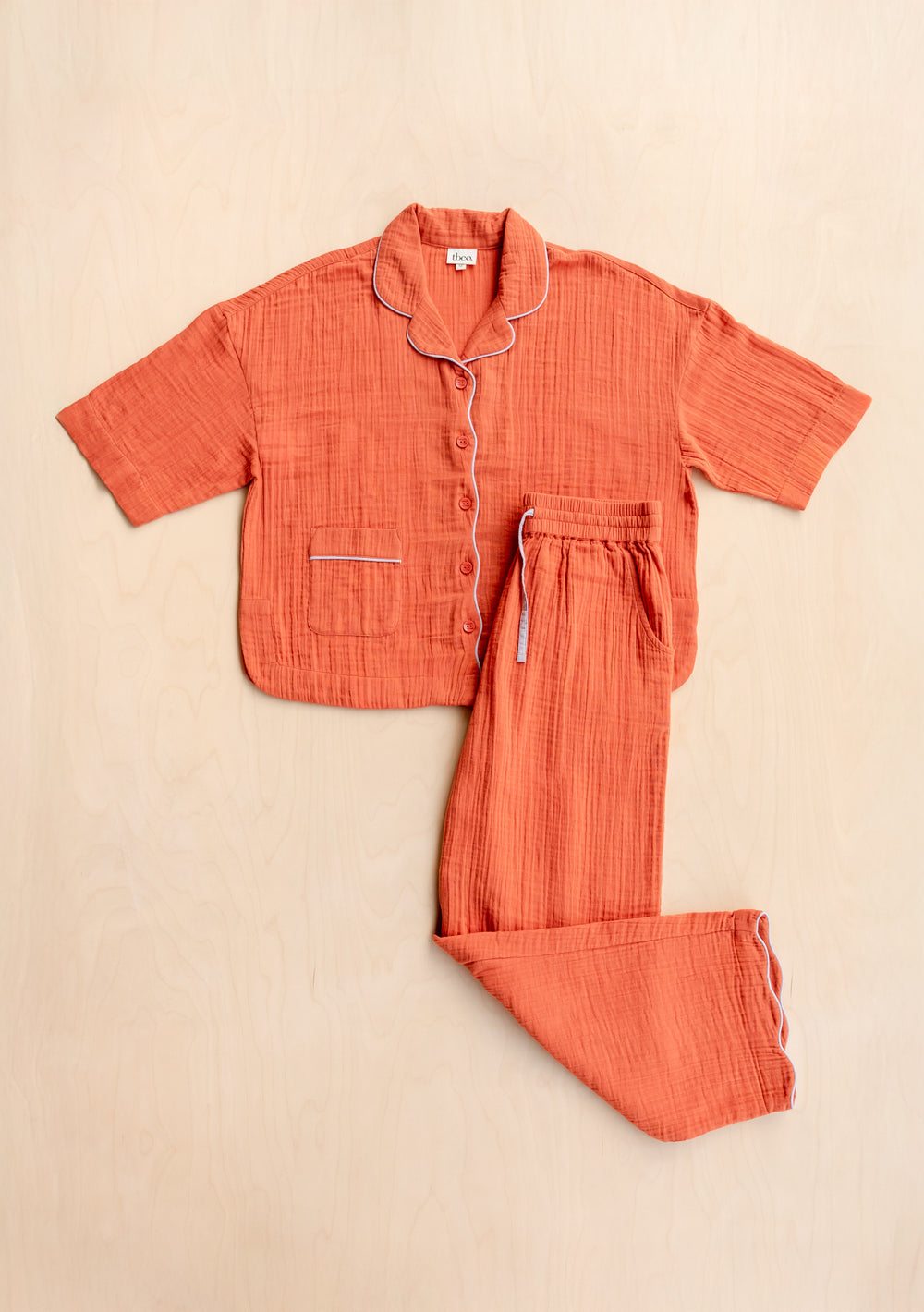 TBCo Cotton Pyjamas in Rust with lilac piping, featuring short sleeve top and relaxed drawstring trousers laid flat on wood surface
