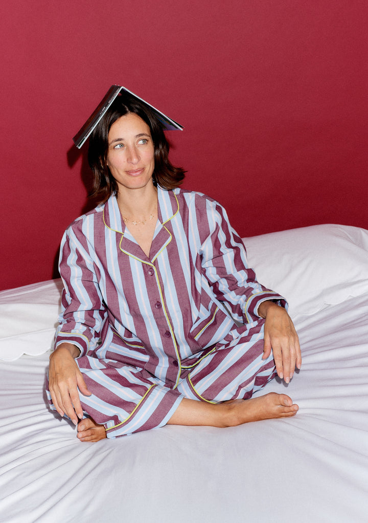 Model in TBCo x Glassette Cotton Pyjamas in Berry Stripe, seated cross-legged on a bed, wearing striped pyjamas with a book on their head, looking amused.