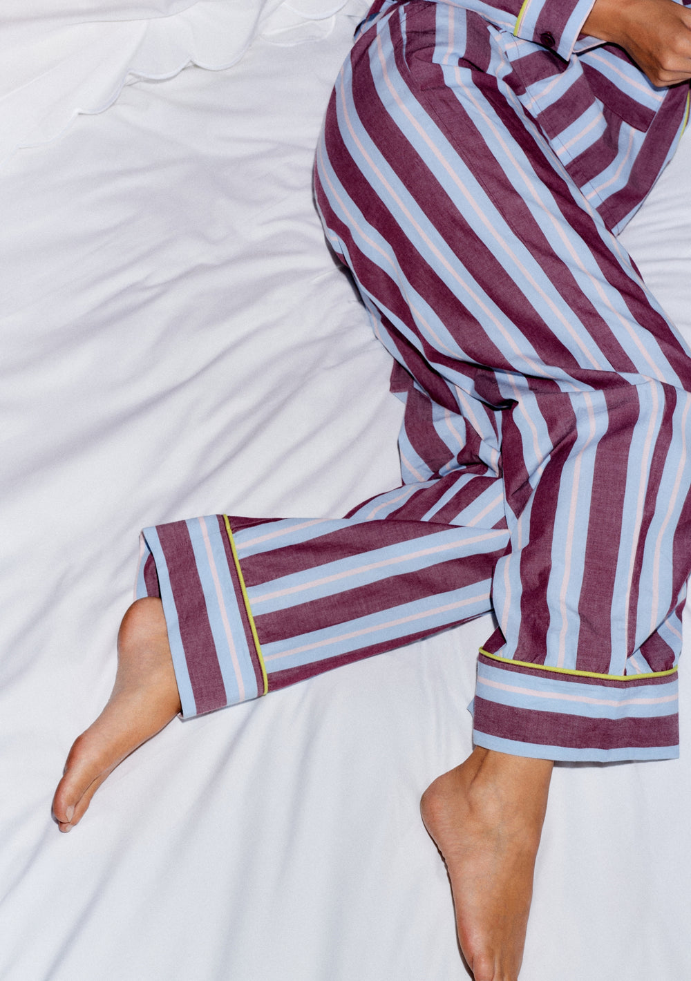 TBCo x Glassette Cotton Pyjamas in Berry Stripe featuring vertical stripes and neon trim, seen on a reclined person with bare feet on a white bed.