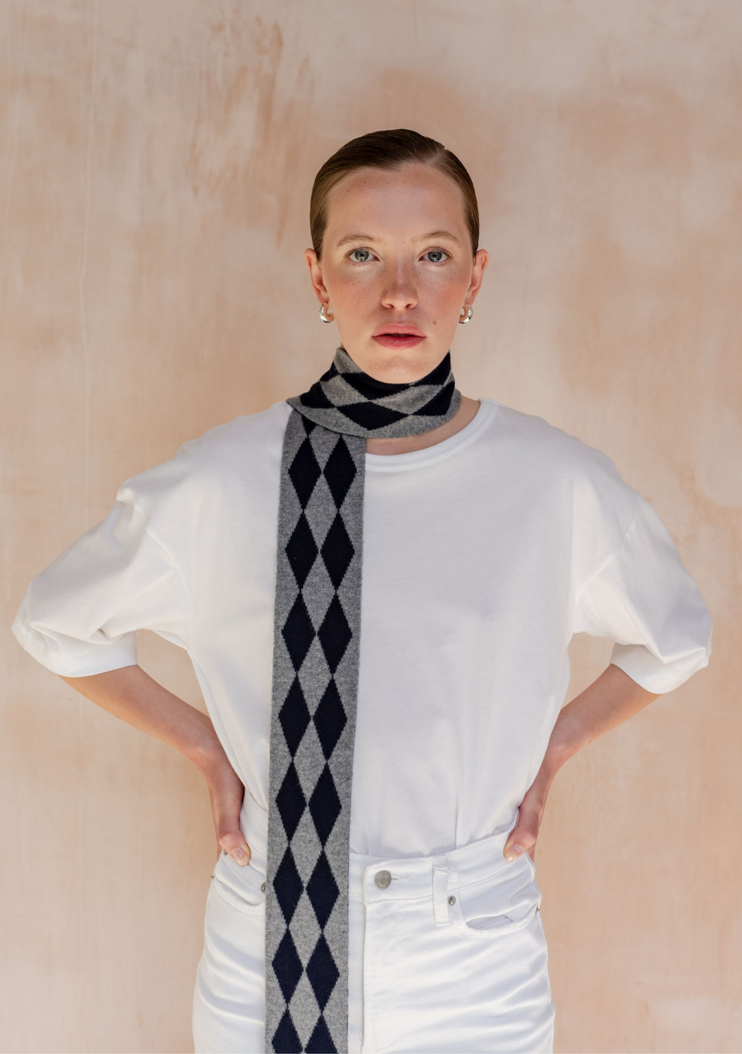 A model wearing TBCo Merino Wool Skinny Scarf in Charcoal Harlequin, featuring grey and dark-navy diamond pattern, over a white T-shirt.