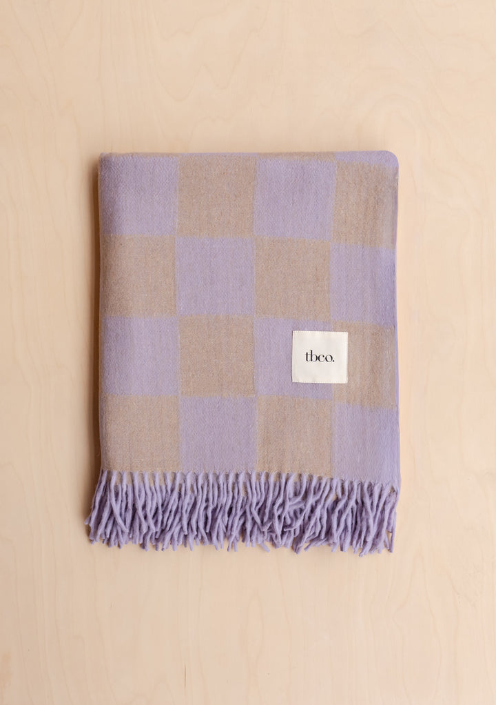 Folded TBCo Recycled Wool Blanket in Lilac & Coffee Checkerboard with fringe detail on light wood background