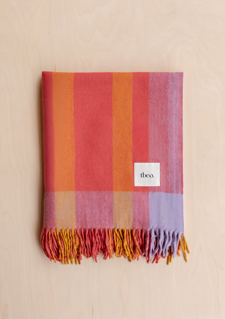 Folded TBCo Recycled Wool Blanket in Orange Frame Stripe with multicoloured fringes on pale wood background