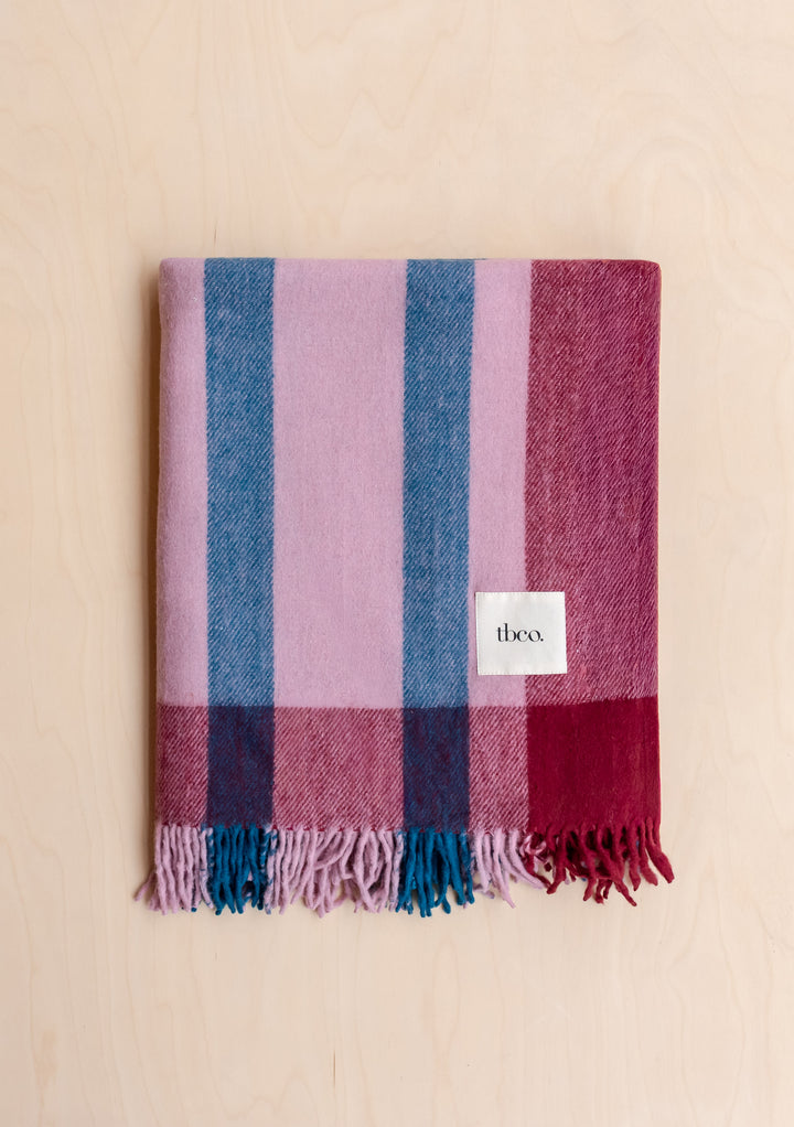 Folded Recycled Wool Blanket in Berry Frame Stripe by TBCo featuring soft fringes and brand label on pale wood surface