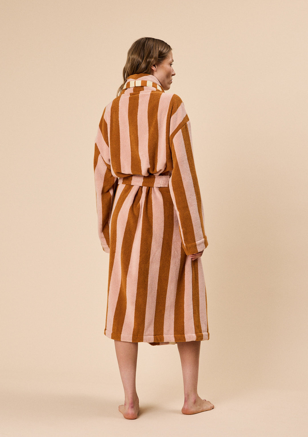 Back view of a woman wearing TBCo Terry Cotton Bath Robe in Honeycomb Stripe with warm brown and beige stripes and belt tied at waist