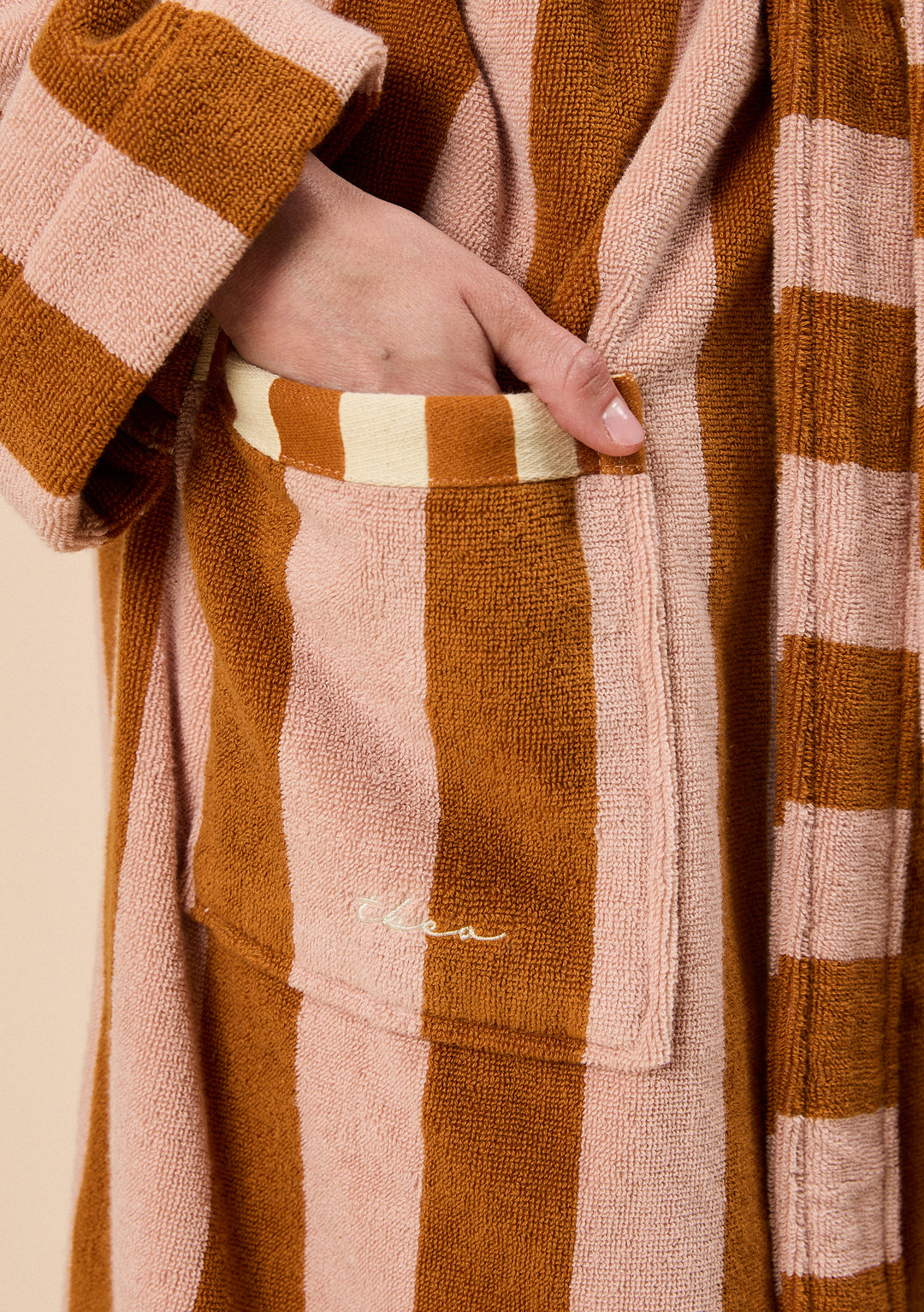 Close-up of a hand in TBCo Terry Cotton Bath Robe in Honeycomb Stripe pocket with soft textured orange and beige stripes