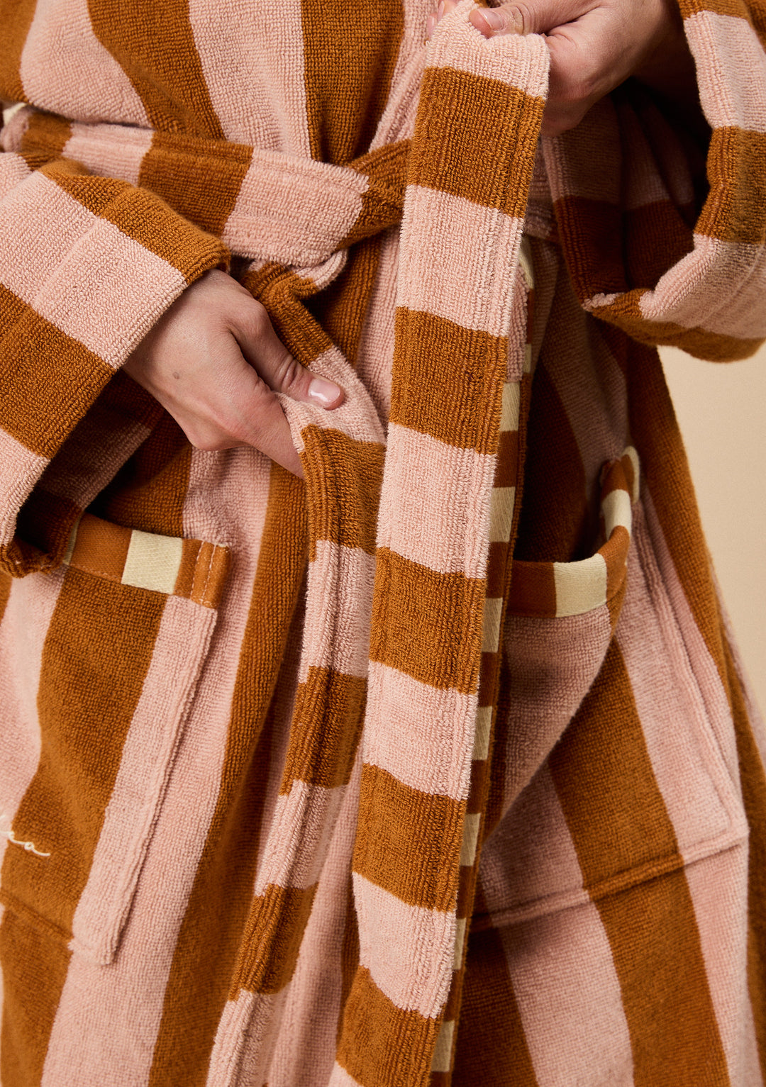 Close-up of hands tying the belt on TBCo Terry Cotton Bath Robe in Honeycomb Stripe with textured warm brown and pink stripes